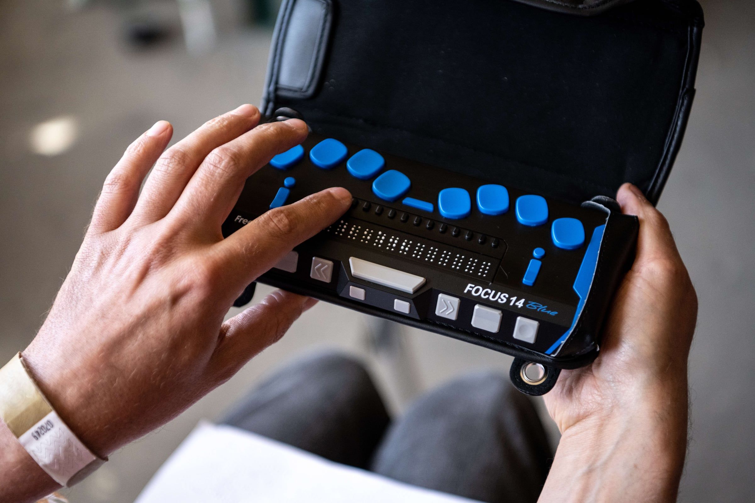 Two hands holding a portable refreshable braille display on their lap, fingertips reading the row of braille dots between the blue control buttons